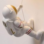 Luminária de astronauta em posição de flutuação, com detalhes em branco e dourado, suspensa por um fio.