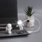 Luz de leitura em forma de astronauta com braço flexível conectada a um portátil, ao lado de uma planta suculenta em vaso de vidro.
