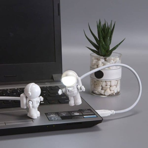 Luz de leitura em forma de astronauta com braço flexível conectada a um portátil, ao lado de uma planta suculenta em vaso de vidro.