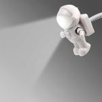 Luminária de astronauta em miniatura com luz LED, conectada por cabo USB, com detalhes brancos e preto, em fundo cinza.