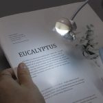 Luz LED portátil em forma de astronauta com braço flexível iluminando uma página de livro sobre eucalipto.