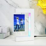 Candeeiro com porta-retratos em levitação, exibindo uma foto de duas pessoas na praia, com iluminação LED colorida na moldura branca, ao lado de livros e flores amarelas.