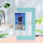 Candeeiro de mesa com estrutura azul em forma de 'C' e porta-retratos digital exibindo uma foto de um casal na praia, com plantas verdes ao redor.