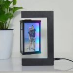 Candeeiro de mesa com estrutura de metal preto e porta-retratos em levitação exibindo uma foto de um casal na praia, com uma planta verde ao lado.