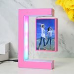 Candeeiro de mesa em forma de letra C com porta-retratos em levitação, iluminação LED azul e rosa, sobre superfície branca com fundo de mármore.