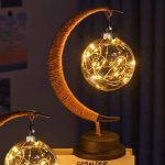 Luminária decorativa com forma de lua, contendo uma bola de vidro com luzes LED internas, apoiada por uma estrutura de madeira curvada.