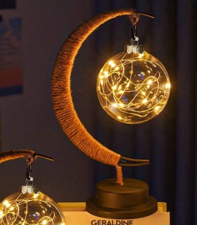 Luminária decorativa com forma de lua, contendo uma bola de vidro com luzes LED internas, apoiada por uma estrutura de madeira curvada.