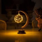 Luminária decorativa com forma de lua crescente e esfera de estrelas brilhantes, em tons dourados, sobre mesa de madeira, ambiente acolhedor e escuro.