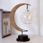Luminária decorativa com forma de lua crescente envolta em corda de sisal, com lâmpadas LED dentro de uma esfera de vidro, apoiada em base preta.