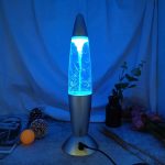 Lâmpada de lava com efeito de tornado azul, exibindo movimento de bolhas e fluxo de líquido, com decoração de plantas e objetos ao fundo.