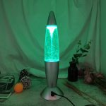 Lâmpada de lava com efeito de tornado em movimento, luz azul e verde, base prateada, ambiente decorativo com plantas e objetos científicos.