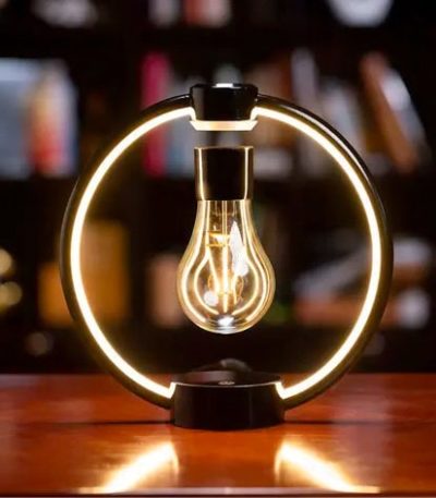 Lâmpada de filamento suspensa dentro de um anel de luz LED circular, com fundo de estantes de livros desfocado.