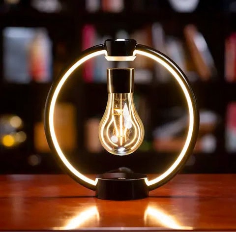 Lâmpada de filamento suspensa dentro de um anel de luz LED circular, com fundo de estantes de livros desfocado.