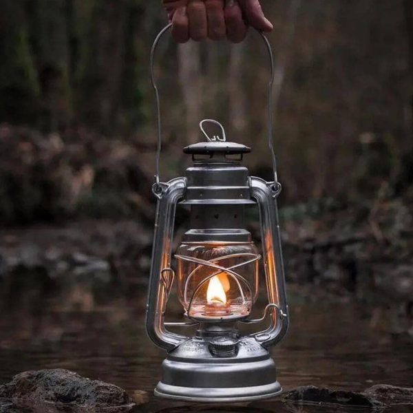 Lanterna a óleo antiga com chama acesa, segurada por uma mão, em cenário de rio ou lago com vegetação ao fundo.