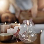 Candeeiro de mesa a óleo com ranhuras de vidro transparente, com vela acesa, rodeado por velas de diferentes tamanhos e cores, numa superfície de madeira.