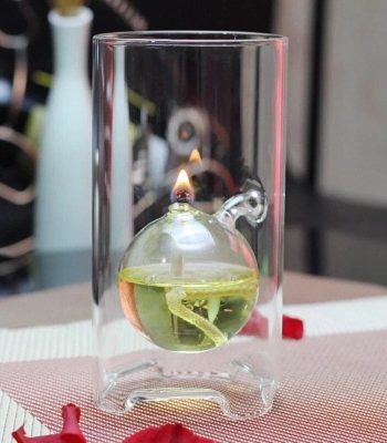 Esfera de vidro com óleo e uma chama acesa no topo, dentro de um tubo de vidro transparente, com fundo desfocado de laboratório.