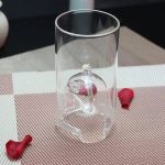 Vaso de vidro transparente com vela acesa no interior, rodeado por pequenos ossos de plástico vermelho.