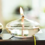 Candeeiro de mesa a óleo com forma de seixo, com chama acesa, feito de vidro transparente, colocado numa superfície escura, com fundo desfocado.