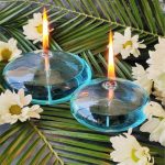 Dois candeeiros de mesa a óleo em forma de seixo, com chama acesa, colocados sobre folhas verdes e flores brancas ao redor.
