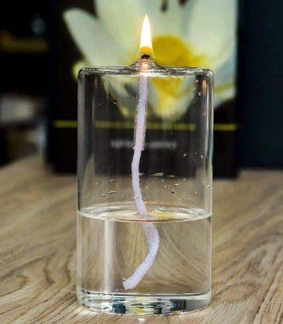 Vela acesa dentro de um vidro transparente com água, com uma flor branca ao fundo.