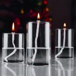 Três velas de vidro com chama acesa, uma maior no centro e duas menores ao lado, com fundo escuro e luz suave refletida na superfície.