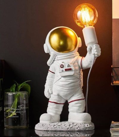 Estatueta de astronauta com capacete dourado a segurar uma lâmpada de plasma acesa, em ambiente de decoração com elementos científicos.