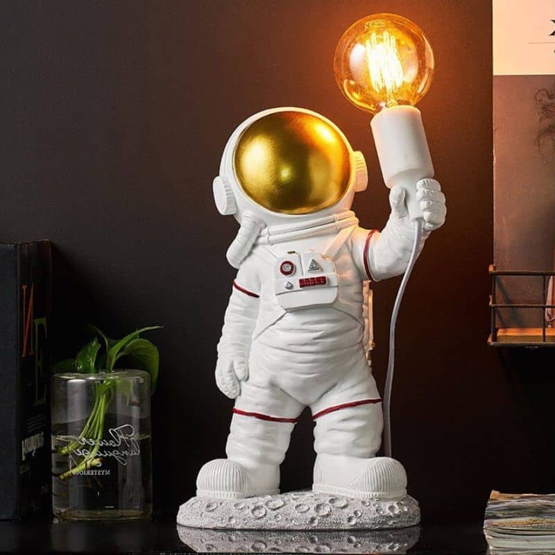 Estatueta de astronauta com capacete dourado a segurar uma lâmpada de plasma acesa, em ambiente de decoração com elementos científicos.