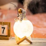 Miniatura de astronauta sentado na lua, segurando uma vara de pesca, com luz quente e base de madeira.