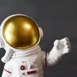 Estátua de astronauta com capacete dourado brilhante, roupa branca com detalhes vermelhos, braço estendido, fundo cinzento escuro.