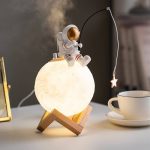 Candeeiro de mesa com forma de lua, astronauta em cima, com detalhes dourados e uma estrela pendurada, ideal para decoração científica infantil.