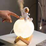 Miniatura de astronauta em posição de exploração na superfície lunar, com luz de lua em uma base de madeira, com estrela pendurada ao lado.