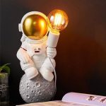 Estátua de astronauta com capacete dourado segurando uma lâmpada acesa, sentado numa base que parece a Lua, decorativo para espaço ou ciência.