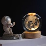 Lâmpada de mesa com design de lua dentro de uma esfera de vidro, acompanhada por um pequeno astronauta de brinquedo em uma base de rocha.