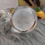 Bola de cristal transparente com detalhes internos, colocada sobre uma superfície de pelúcia cinzenta, com pequenos objetos decorativos ao fundo.