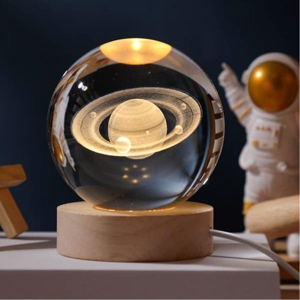 Lâmpada de mesa com design de planeta com anéis, iluminação suave, base de madeira clara, fundo escuro, decoração científica moderna.