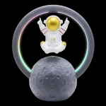 Miniatura de astronauta com capacete dourado, rodeado por anel de luz LED colorida, apoiado numa réplica da Lua com textura lunar detalhada.