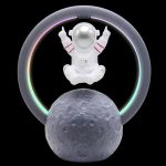 Miniatura de astronauta com capacete prateado, sentado sobre uma lua artificial, rodeado por anel de luz LED multicolorido, fundo preto.
