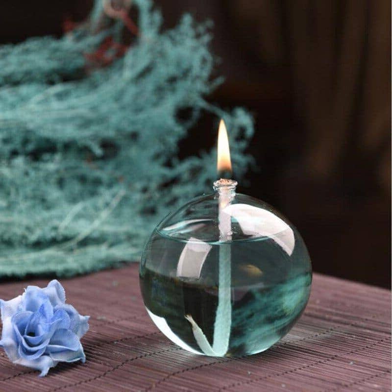 Candeeiro de mesa redondo a óleo com chama acesa, feito de vidro transparente, colocado numa superfície de madeira, com decoração de tecido azul ao lado.