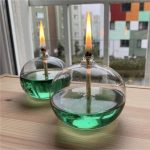 Candeeiro de mesa com forma de esfera de vidro, contendo líquido verde e uma vela acesa no centro, colocado numa superfície de madeira junto a uma janela.
