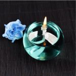 Candeeiro de mesa redondo a óleo com chama acesa, colocado sobre uma superfície escura com uma rosa azul ao lado.
