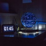 Lâmpada de mesa com esfera que projeta constelações azuis, iluminada em ambiente escuro, com relógio digital ao lado.