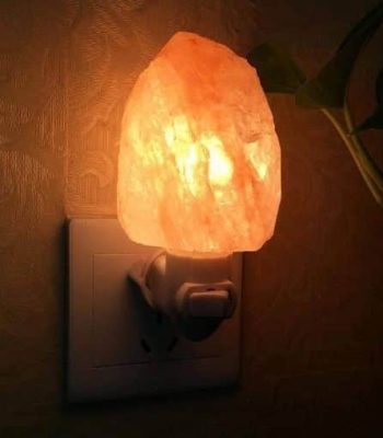 Candeeiro de sal em forma de pedra, iluminado, ligado a uma tomada na parede, com uma planta ao lado.