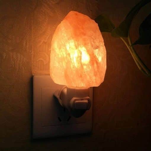 Candeeiro de sal em forma de pedra, iluminado, ligado a uma tomada na parede, com uma planta ao lado.