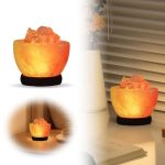 Lâmpada de sal em forma de rosa, iluminada, com base preta, colocada numa mesa ao lado de uma vela e uma planta.