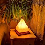 Lâmpada de sal em forma de pirâmide iluminada, colocada numa mesa ao lado de uma planta e cortinas de renda.