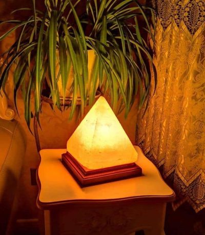 Lâmpada de sal em forma de pirâmide iluminada, colocada numa mesa ao lado de uma planta e cortinas de renda.