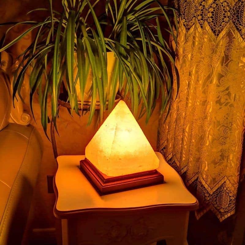 Lâmpada de sal em forma de pirâmide iluminada, colocada numa mesa ao lado de uma planta e cortinas de renda.