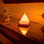 Candeeiro de sal em forma de pirâmide, iluminado com luz quente, colocado numa superfície de mármore, com decoração de árvore de Natal ao fundo.