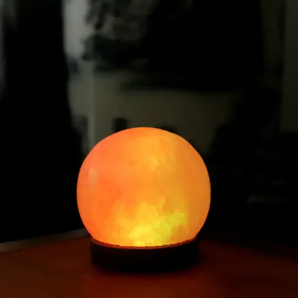 Candeeiro de sal redondo iluminado, com luz quente, colocado sobre uma superfície de madeira, ambiente escuro ao fundo.