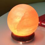 Candeeiro de sal redondo iluminado com luz quente, colocado numa base de madeira escura, numa mesa branca com livros vermelhos ao fundo.